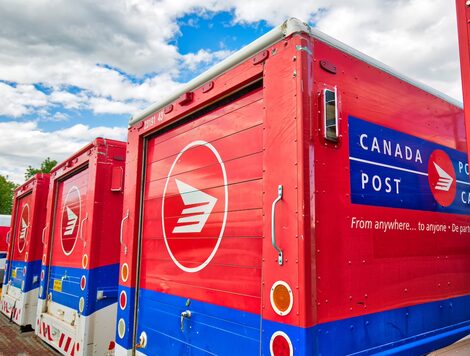 A row of red, blue and white striped mail trucks, with "Canada Post" written in white lettering on a blue square across the side of each vehicle