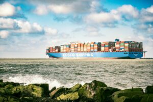 A CONTAINER SHIP BEARING THE WORD COSCO IN GIANT WHITE LETTERS PLIES THE OCEAN, WITH ROCKS IN THE FOREGROUND