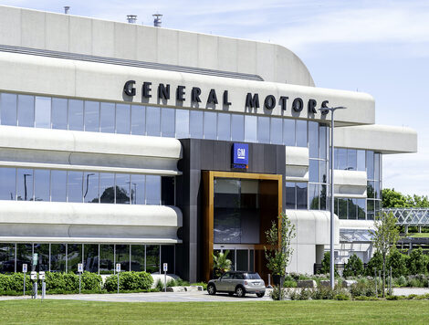 THE EXTERIOR OF A GM TECHNICAL CENTER CAMPUS CAN BE SEEN DURING A SUNNY DAY. A CAR IS PARKED RIGHT NEAR THE FACILITY'S FRONT ENTRANCE. 