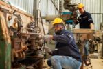 manufacturing-factory-iStock-1459502685.jpg A man wearing a blue long-sleeve shirt and jeans, with a yellow hard hat, kneeling down in front of a factory machine, with a similarly dressed man standing behind him in the background.
