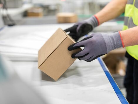 A PERSON WEARING GLOVES AND A HI-VIS VEST INSPECTS A BROWN CARDBOARD BOX ON A CONVEYOR SYSTEM
